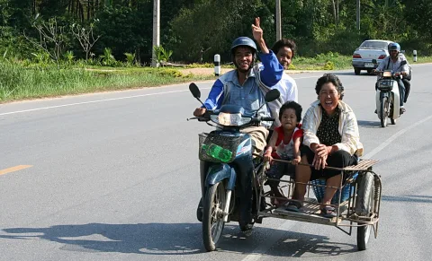 Ein Mann mit Helm fährt einen Motorroller mit Seitenwagen über eine Landstraße. Auf dem Roller sitzen ein Mann, der die Hand zum Victory-Zeichen hebt, ein kleines Kind und eine lachende Frau im Beiwagen.