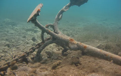Rostiger Anker mit Kette auf steinigem Meeresgrund, im Hintergrund Seegras unter türkisfarbenem Wasser.