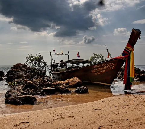 Ein hölzernes Langboot mit bunten Bändern am Bug liegt auf einem Sandstrand neben dunklen Felsen, darüber ziehen dunkle Wolken auf.