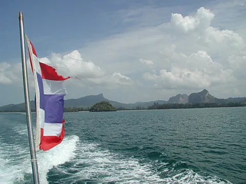 Thailändische Flagge an einem Bootsheck, dahinter Meer, bewaldete Insel und felsige Kalksteinberge unter Wolken