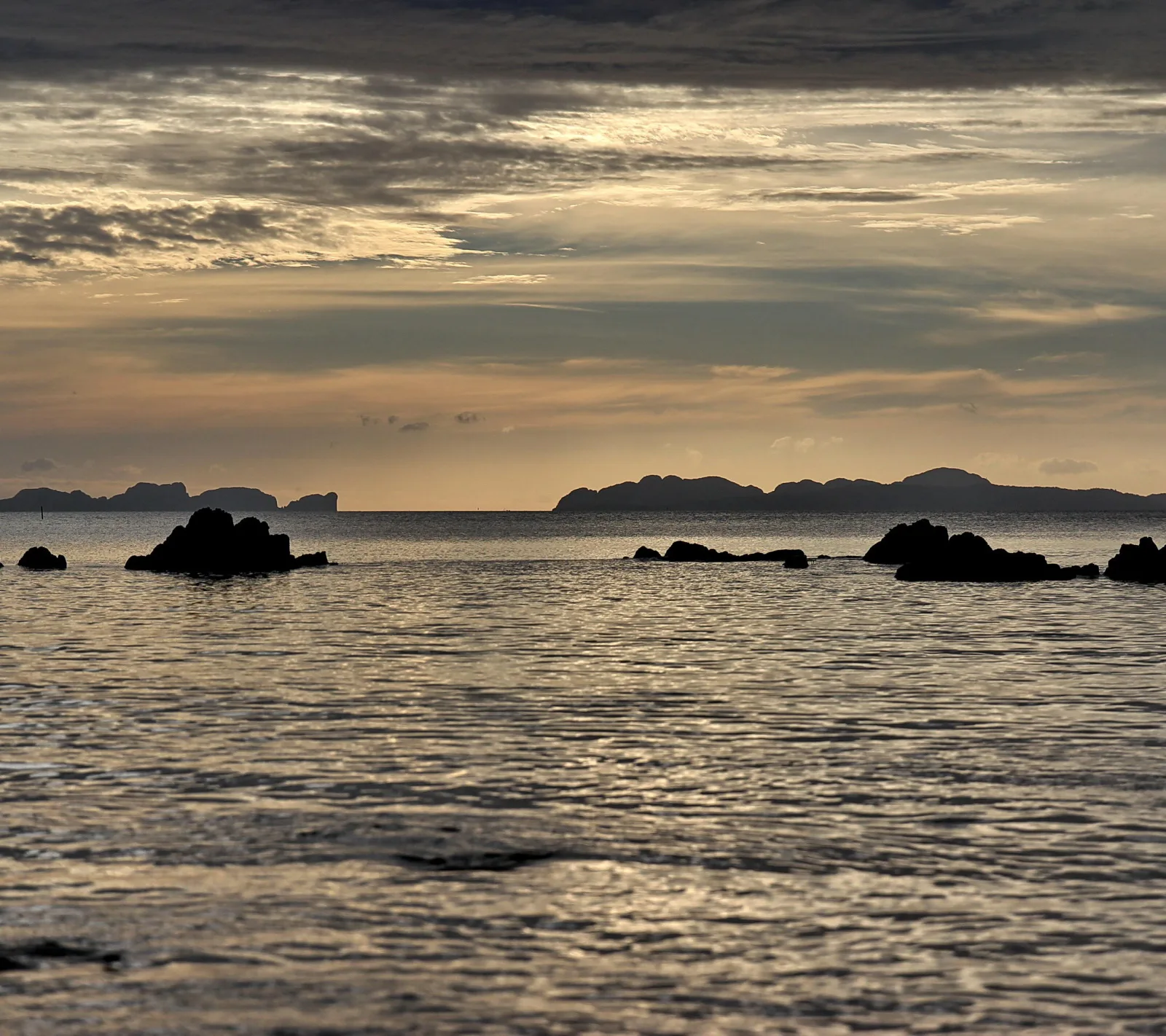 Ruhige Meeresoberfläche am Abend mit dunklen Felsen im Wasser und Inselsilhouetten am Horizont unter einem bewölkten, golden gefärbten Himmel.
