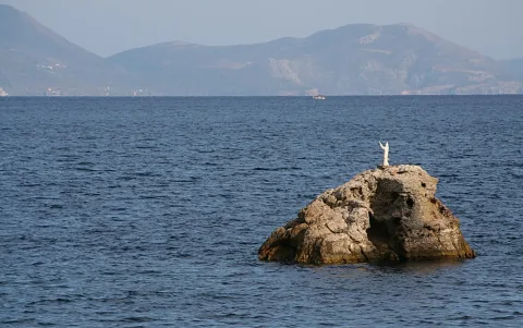 Eine weiße Jesusstatue mit erhobenen Armen steht auf einem zerklüfteten Felsen im tiefblauen Meer, im Hintergrund liegen hazy Berge.
