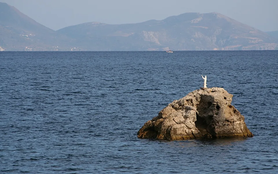 Eine weiße Jesusstatue mit erhobenen Armen steht auf einem zerklüfteten Felsen im tiefblauen Meer, im Hintergrund liegen hazy Berge.