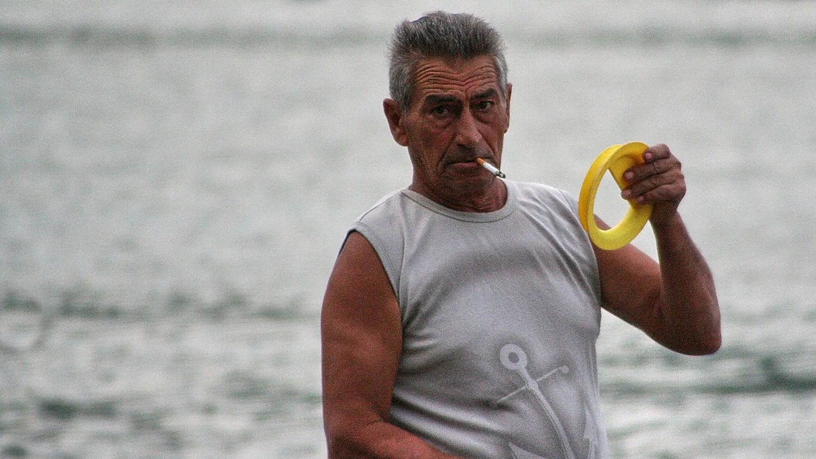 Älterer Mann mit grauen Haaren, ärmellosem Shirt mit Ankermotiv und Zigarette im Mund hält einen gelben Ring vor einem unscharfen Meer.