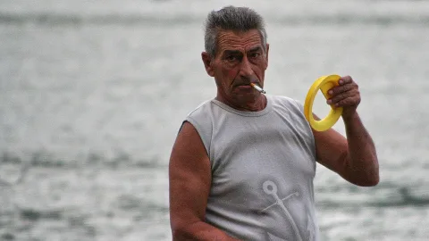 Älterer Mann mit grauen Haaren, ärmellosem Shirt mit Ankermotiv und Zigarette im Mund hält einen gelben Ring vor einem unscharfen Meer.