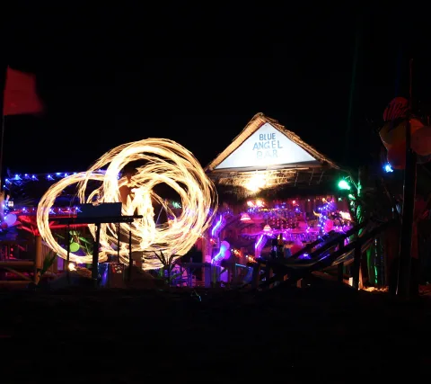 Nachtaufnahme einer Strandbar mit Strohdach und Schild 'Blue Angel Bar'. Links kreist eine Feuershow als lange, leuchtende Lichtspur, rechts sind bunte Lichterketten und Luftballons zu sehen.