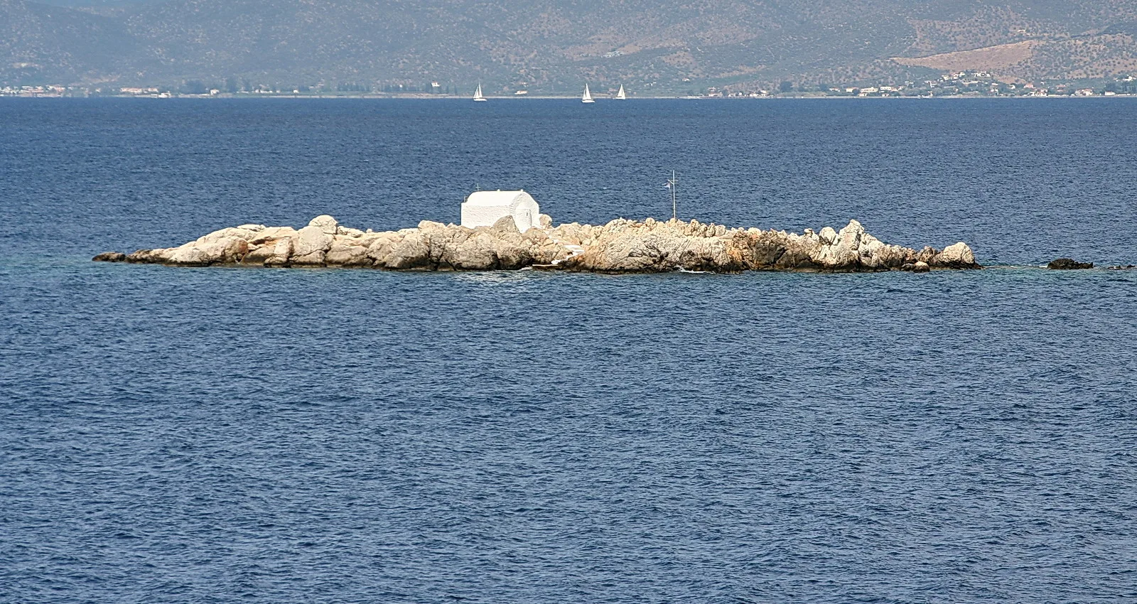Felsige Insel mitten im blauen Meer, darauf eine weiße kleine Kapelle und ein dünner Mast; im Hintergrund eine bergige Küste mit Häusern und kleinen Segelbooten.