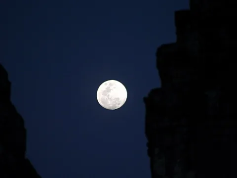 Heller Vollmond am dunkelblauen Nachthimmel, eingerahmt von zwei dunklen Steinmauern.
