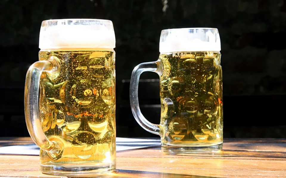 Zwei gefüllte Biergläser mit Henkel stehen auf einem Holztisch vor dunklem Hintergrund, Sonnenlicht fällt auf den Schaum.