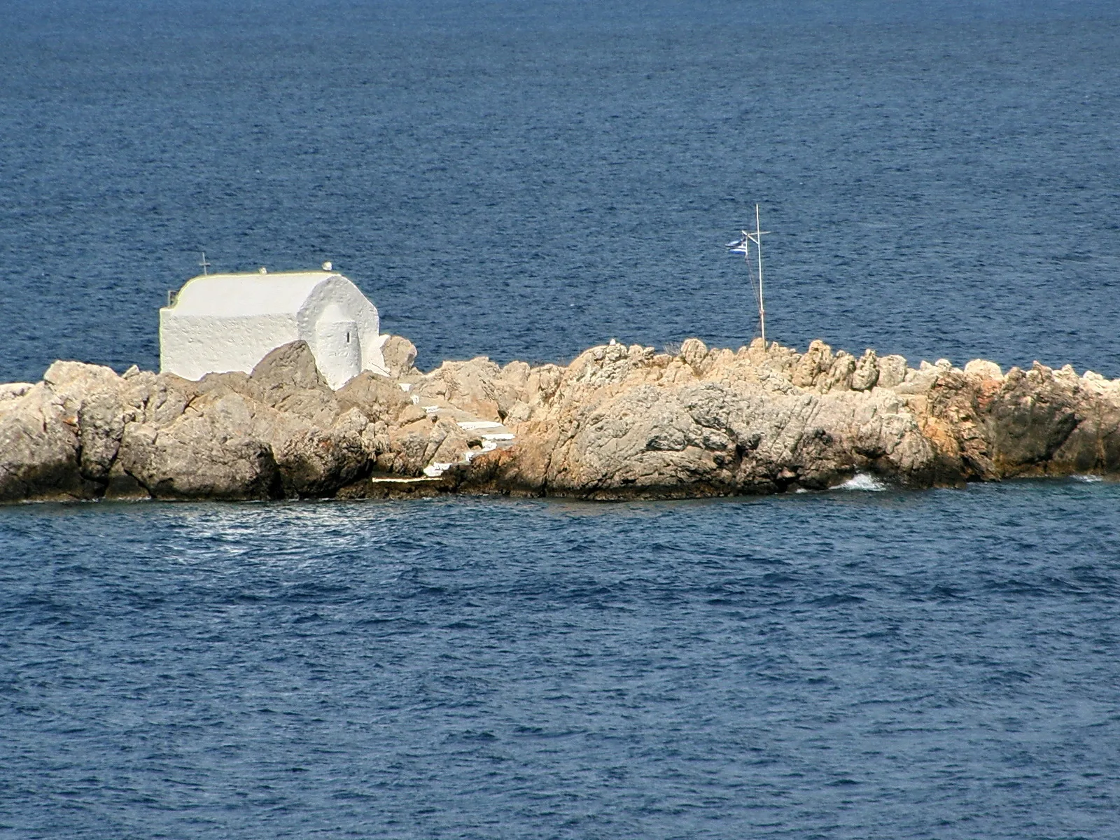 Eine weiß getünchte kleine Kapelle steht auf einem felsigen Vorsprung im Meer, daneben ragt ein Mast mit einer griechischen Flagge auf.