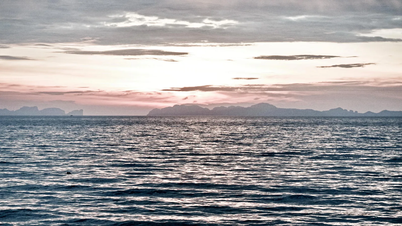 Ruhige Meeresoberfläche mit leichten Wellen, im Hintergrund eine Küstenkette aus Bergen und Inseln unter einem Himmel mit Wolken und rosa Schimmer am Horizont.