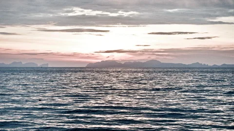 Ruhige Meeresoberfläche mit leichten Wellen, im Hintergrund eine Küstenkette aus Bergen und Inseln unter einem Himmel mit Wolken und rosa Schimmer am Horizont.