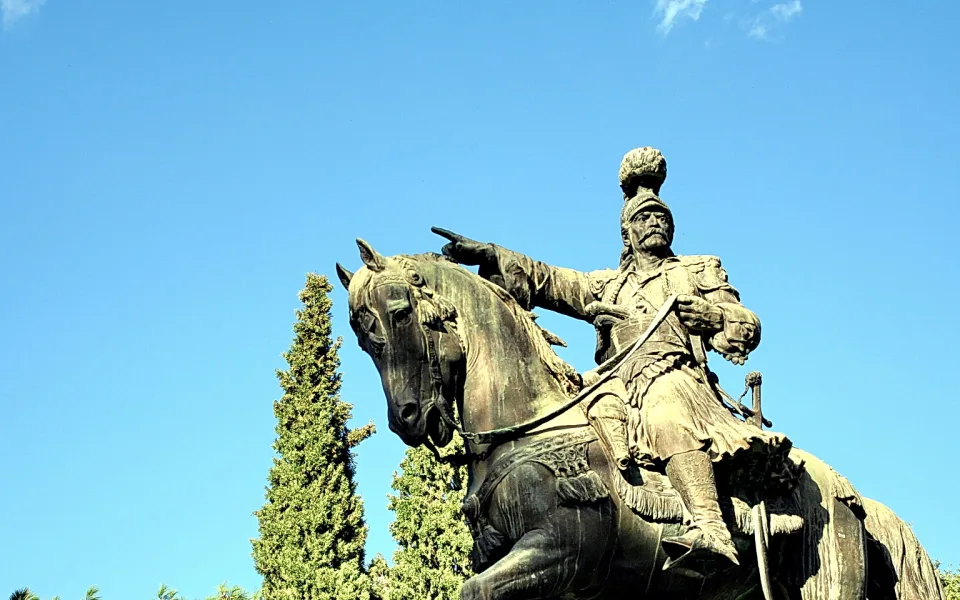 Bronzestatue eines Reiters auf einem Pferd, der mit ausgestrecktem Arm nach links zeigt, vor wolkenlosem blauem Himmel und Zypressen.