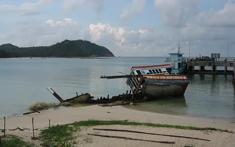 Ein zerfallenes buntes Holzboot liegt schräg auf dem Sand einer Bucht, links davon ein halb versunkenes Wrack, rechts ein Steg mit Anlegestelle und ein Passagierboot vor bewaldetem Hügel unter bewölktem Himmel.