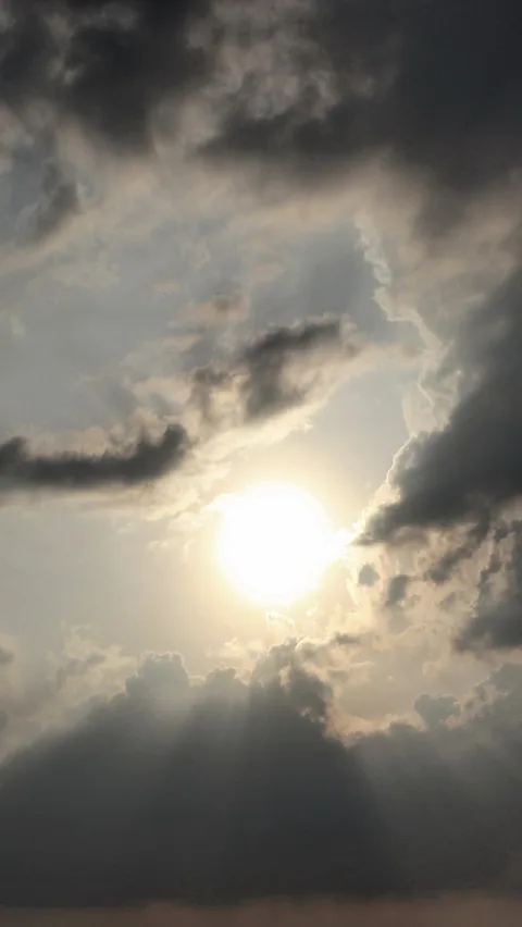 Die Sonne scheint durch eine Lücke in einer Wolkendecke, darunter und daneben dunkle Wolkenbänke, der Himmel ist grau-weiß.