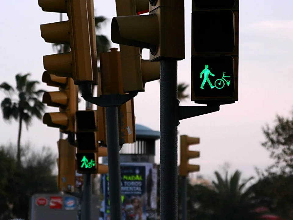 Mehrere gelbe Ampelmasten an einer Straße, an einem Masten leuchten grüne Symbole für Fußgänger und Fahrrad, im Hintergrund Palmen und Dämmerungshimmel.