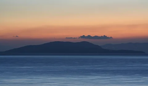 Dunkle Silhouette einer Insel über ruhigem Meer, darüber ein orange-rosa Himmel mit einzelnen Wolken im Morgenlicht.