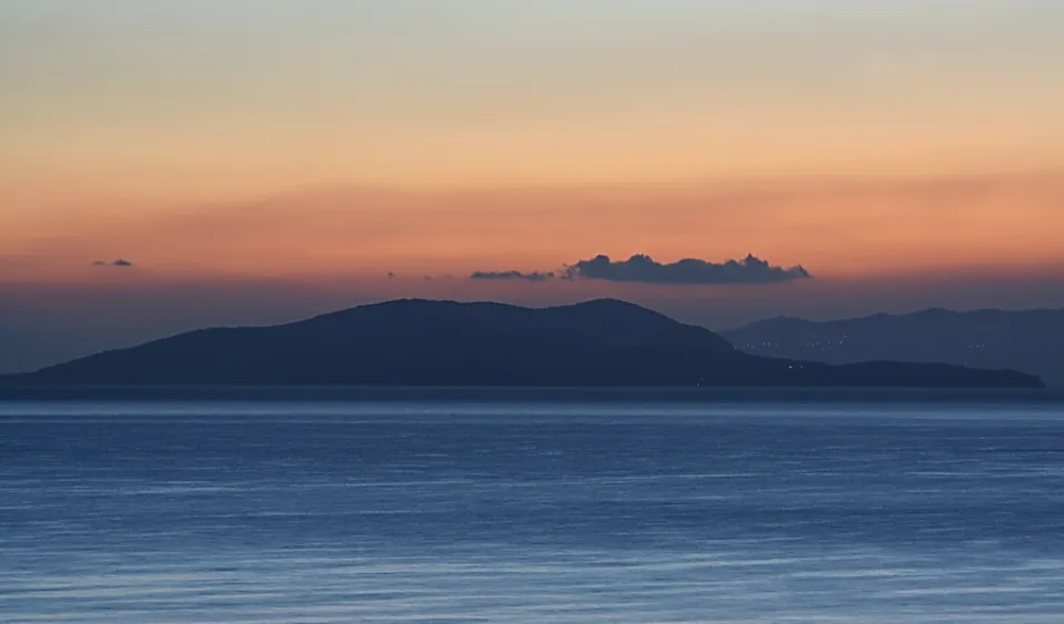 Dunkle Silhouette einer Insel über ruhigem Meer, darüber ein orange-rosa Himmel mit einzelnen Wolken im Morgenlicht.