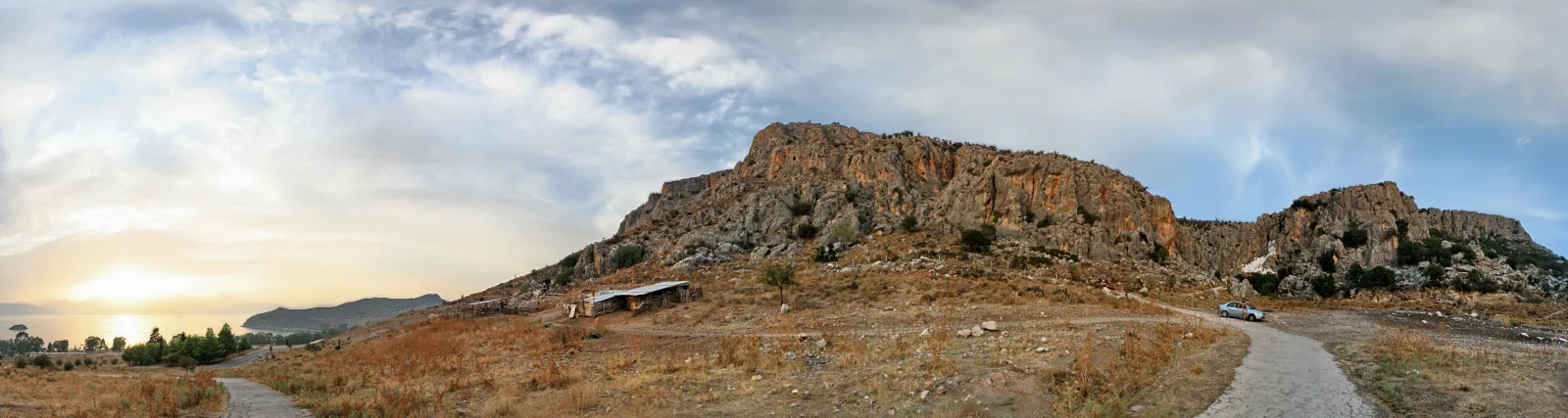 Breites Panorama eines zerklüfteten Felsmassivs mit trockenem Grasland im Vordergrund, einem kleinen Schuppen, einem Feldweg mit parkendem Auto und einem tiefstehenden Sonnenuntergang über dem Meer links.
