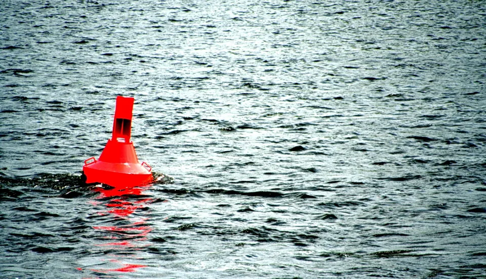 Eine rote Boje schwimmt auf grauem, gekräuseltem Wasser und spiegelt sich leicht im Wasser.