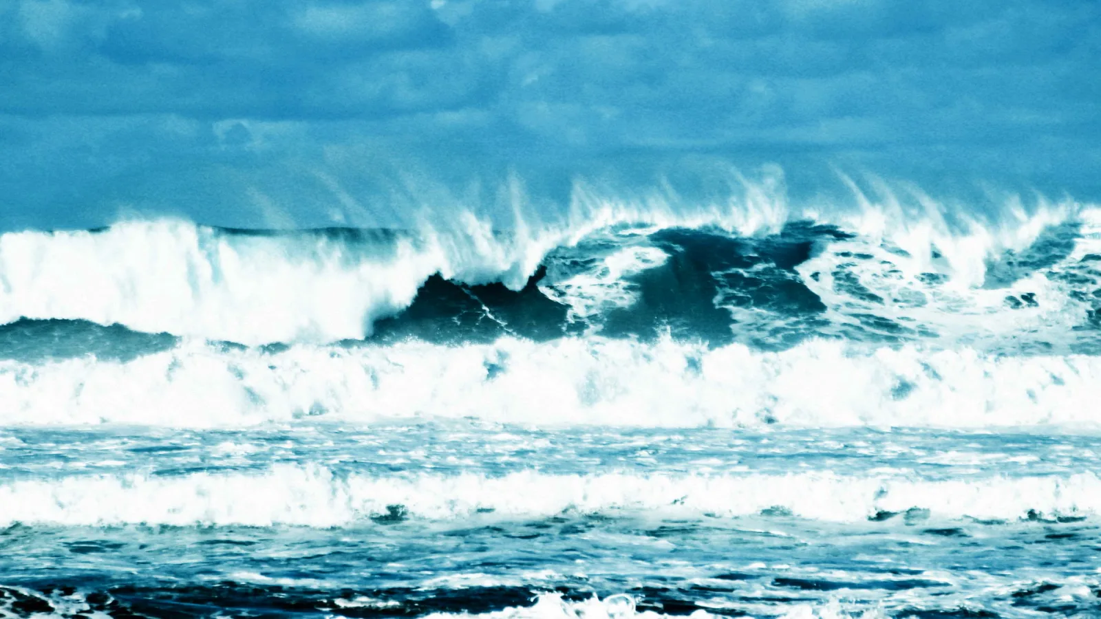 Hohe Wellen brechen sich unter einem bewölkten blaugrauen Himmel auf dem Meer.