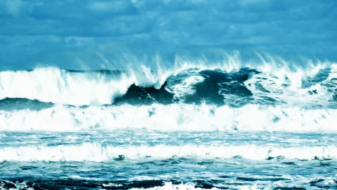 Hohe Wellen brechen sich unter einem bewölkten blaugrauen Himmel auf dem Meer.