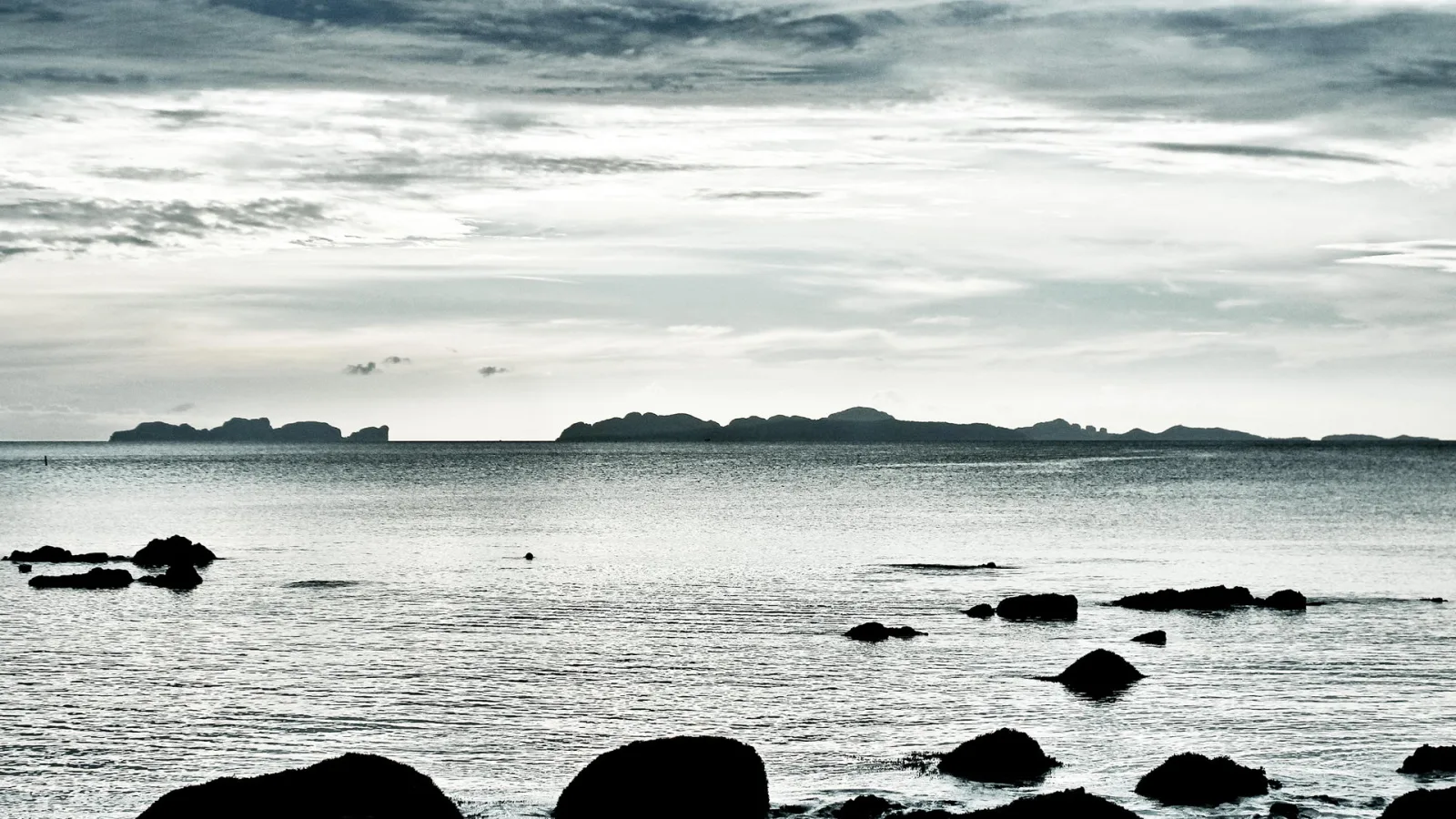 Graues Meer mit dunklen Felsen im Vordergrund, dahinter eine Inselkette am Horizont unter bewölktem Himmel.