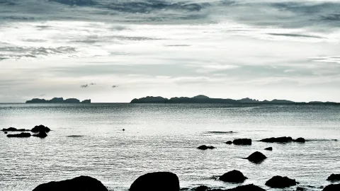 Graues Meer mit dunklen Felsen im Vordergrund, dahinter eine Inselkette am Horizont unter bewölktem Himmel.