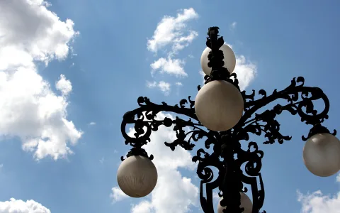Schwarze, verzierte Straßenlaterne mit drei milchweißen Kugelleuchten vor blauem Himmel mit Wolken, von unten fotografiert.