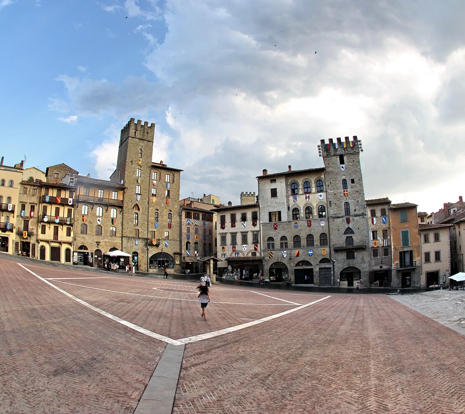 Weiter Backsteinplatz, gesäumt von mittelalterlichen Häusern mit Wappen und Türmen, darüber bewölkter Himmel. Eine einzelne Person geht über den Platz.