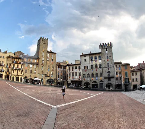 Weiter Backsteinplatz, gesäumt von mittelalterlichen Häusern mit Wappen und Türmen, darüber bewölkter Himmel. Eine einzelne Person geht über den Platz.