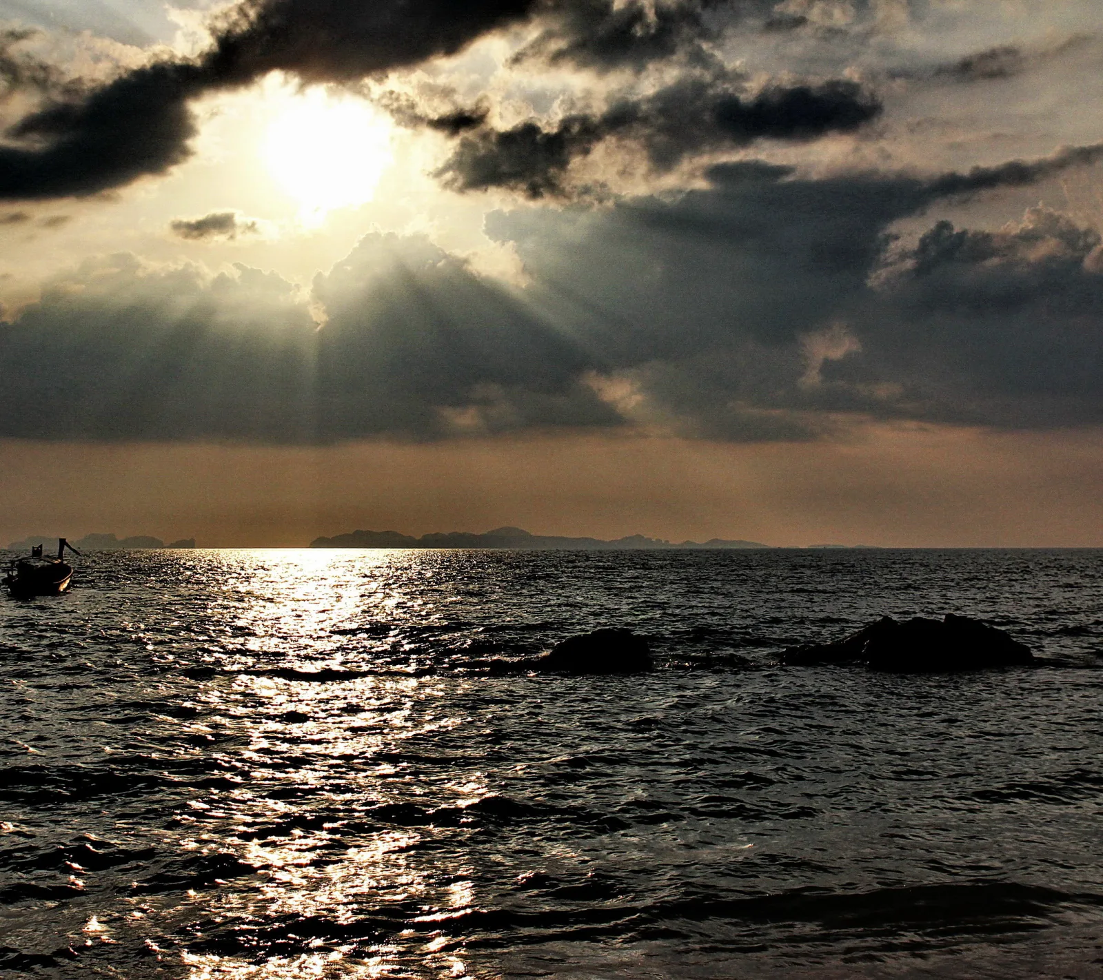Sonne bricht durch dunkle Wolken über einem Meer, rechts im Bild liegen zwei Felsen im Wasser, links treibt ein kleines Boot.