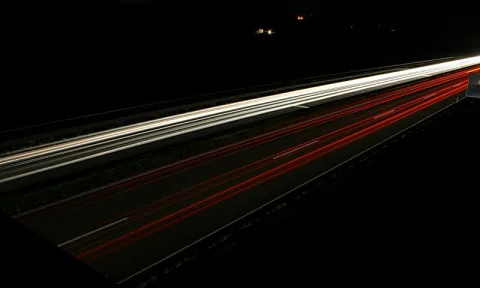 Langzeitbelichtung einer Autobahn bei Nacht: weiße Scheinwerferspuren links, rote Rücklichter rechts, schwarzer Hintergrund.