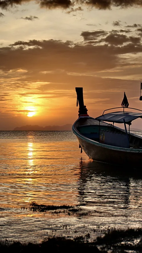 Ein hölzernes Langheckboot liegt am flachen Ufer, rechts im Bild, vor einem Meer mit Sonnenuntergang. Die Sonne steht tief am Horizont, der Himmel ist orange und grau bewölkt, im Wasser spiegeln sich die Lichtbahnen.