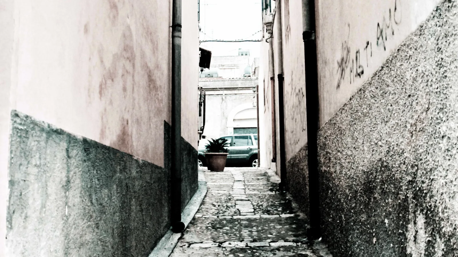 Schmale Gasse mit Kopfsteinpflaster zwischen verwitterten Hauswänden, im Hintergrund ein grünes Auto und ein Blumentopf mit Pflanze.