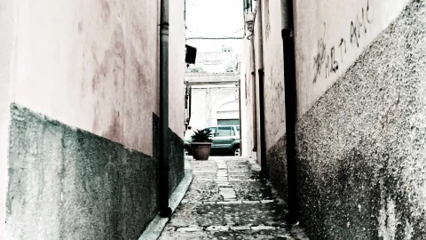 Schmale Gasse mit Kopfsteinpflaster zwischen verwitterten Hauswänden, im Hintergrund ein grünes Auto und ein Blumentopf mit Pflanze.