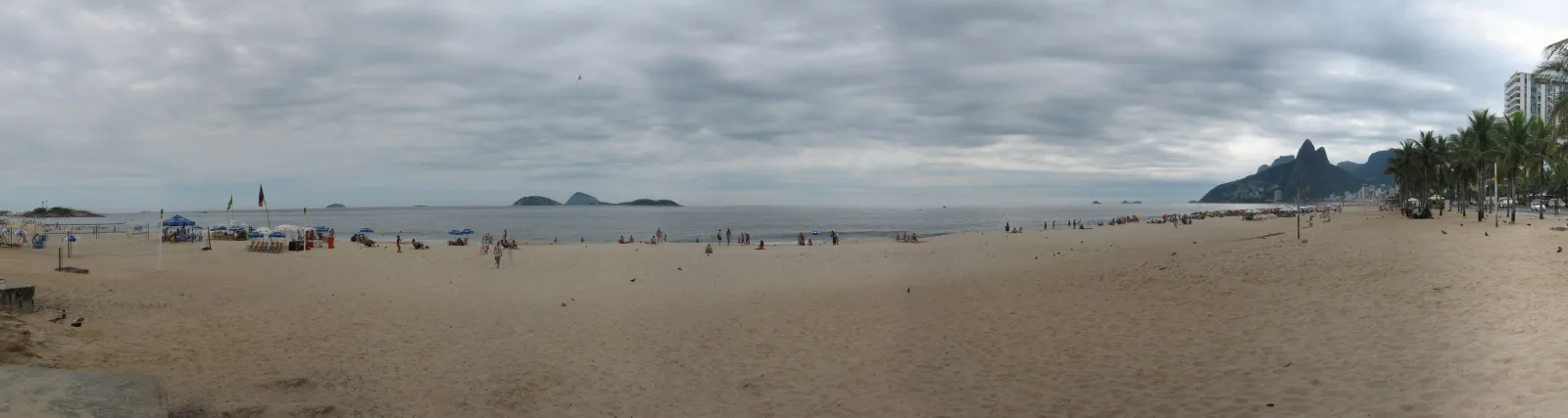 Breiter Sandstrand unter bewölktem Himmel mit wenigen Menschen, im Hintergrund Meer, kleine Inseln und ein bewaldeter Felsberg mit Hochhäusern am rechten Rand.
