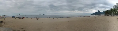 Breiter Sandstrand unter bewölktem Himmel mit wenigen Menschen, im Hintergrund Meer, kleine Inseln und ein bewaldeter Felsberg mit Hochhäusern am rechten Rand.