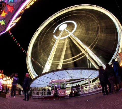 Riesenrad mit weißen Lichterkränzen, im Fisheye-Blick und mit Bewegungsunschärfe aufgenommen, davor Menschen auf einer beleuchteten Plattform und dunkler Nachthimmel