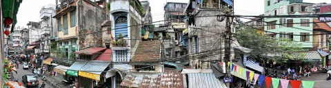 Panoramablick auf eine enge Straße in Hanoi mit verwitterten Altbauten, Stromleitungen, Motorrollern, Marktständen und bunten Wimpelketten.