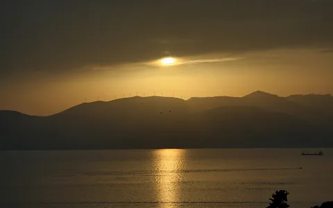 Sonnenuntergang hinter einer Bergkette am Meer, auf dem Bergkamm stehen Windkraftanlagen, rechts ein kleines Schiff auf dem Wasser, unten rechts dunkle Baumsilhouette.