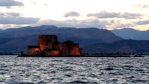 Beleuchtete Festung auf einer kleinen Insel im Meer, dahinter Bergketten bei Abenddämmerung.