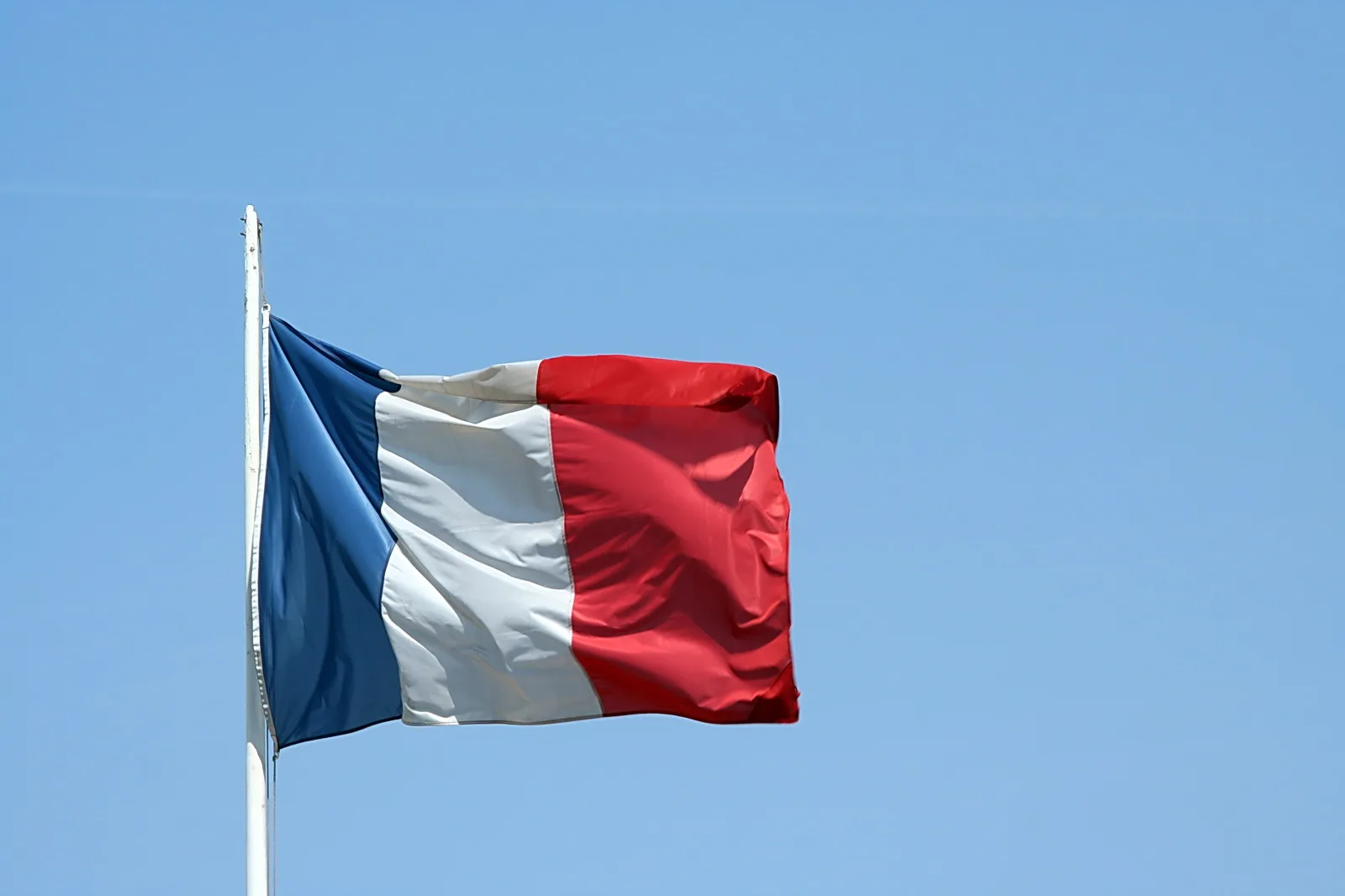 Französische Flagge mit blauem, weißem und rotem Streifen weht an einem weißen Fahnenmast vor blauem Himmel.