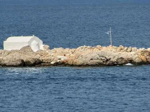 Eine weiß getünchte Kapelle mit gewölbtem Dach steht auf einem felsigen Küstenvorsprung vor dunkelblauem Meer, rechts ragt ein Mast mit griechischer Flagge auf.