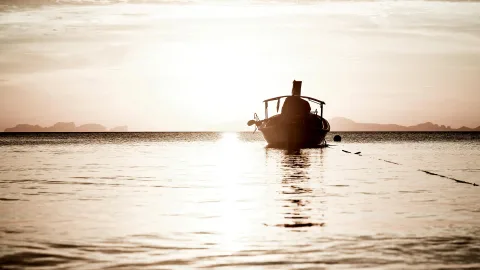 Silhouette eines kleinen Boots auf ruhigem Meer bei Sonnenuntergang, am Horizont Inseln im Dunst.