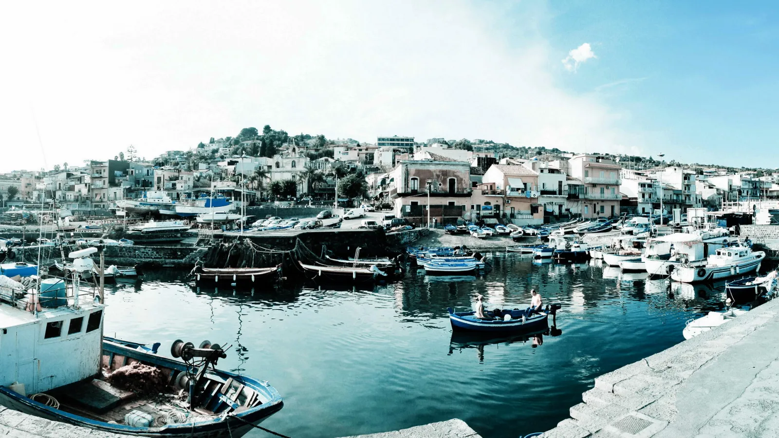 Panoramablick auf einen kleinen Hafen mit blauen Fischerbooten, Booten am Kai und weißen Häusern an einem Hang unter hellem Himmel.