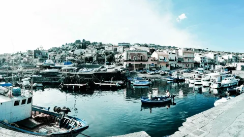 Panoramablick auf einen kleinen Hafen mit blauen Fischerbooten, Booten am Kai und weißen Häusern an einem Hang unter hellem Himmel.