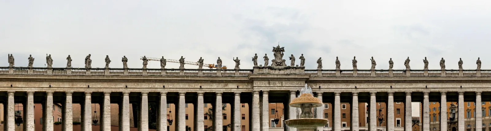 Lange Kolonnade aus hellen Säulen mit einer Reihe von Heiligenstatuen auf der Balustrade, in der Mitte ein Brunnen mit Obelisk-ähnlichem Aufsatz, dahinter Gebäude unter bewölktem Himmel.