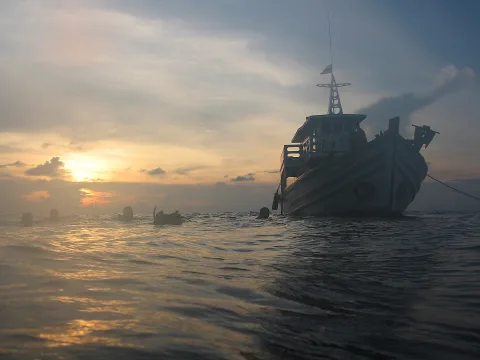 Mehrere Taucher schwimmen an der Wasseroberfläche neben einem Holzboot, im Gegenlicht einer untergehenden Sonne mit bewölktem Himmel.