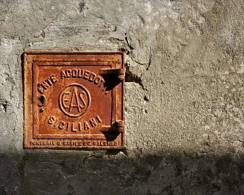 Quadratische, rostbraune Gusseisenklappe mit erhabener Inschrift EAS und Schriftzug Acquedotto Siciliani auf einer verwitterten grauen Putzwand, von links beleuchtet.
